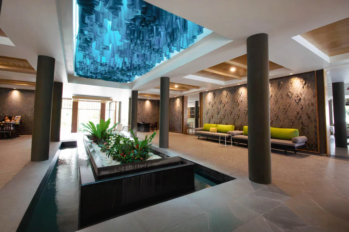 Spacious lobby at Andaman Cannacia Resort in Phuket, featuring a central indoor water garden, modern seating area, and an artistic blue ceiling installation