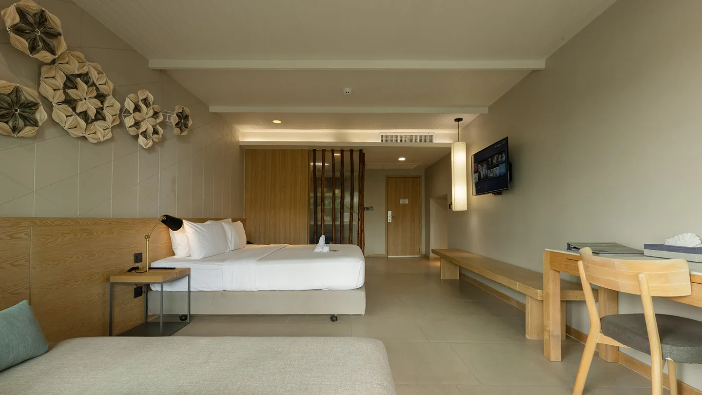 A large double bed with a patterned headboard, a cozy sofa, a wooden desk, and bright, airy décor accented by local artistry and warm lighting