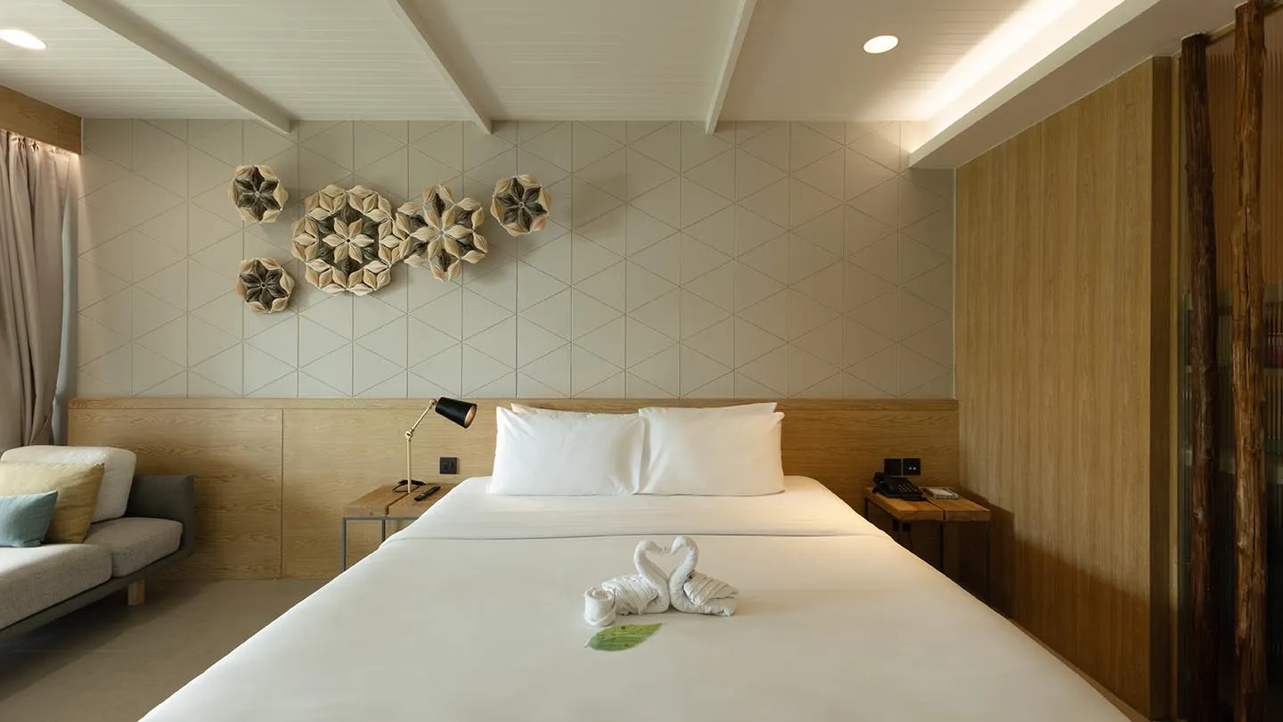 A large bed with swan towels, wooden bedside tables with lamps, a sofa with cushions, and light wood décor accented by a floral-patterned wall art for a serene, airy ambiance