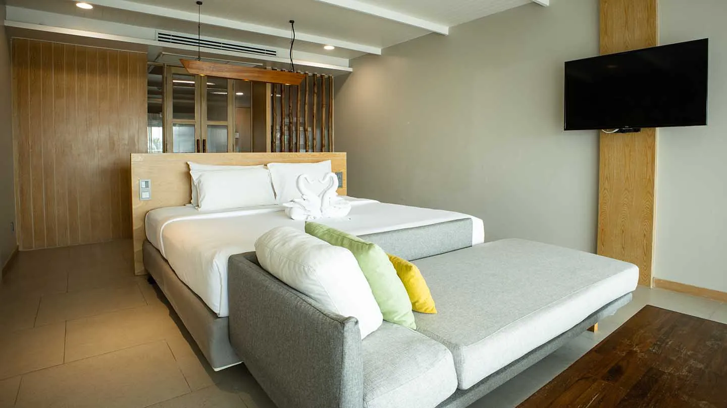 The Canna Deluxe bedroom features a double bed with wooden accents, a cozy sofa with colorful cushions, modern amenities, and floor-to-ceiling windows creating a calm, luxurious ambiance