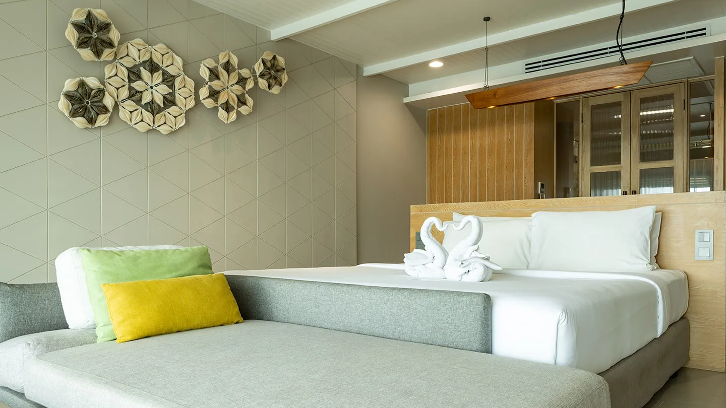 The Canna Deluxe room features a large bed with white linens and a swan towel arrangement, a cozy daybed with green and yellow pillows, and warm, modern décor with subtle Thai accents