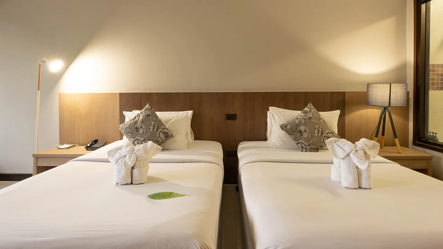A bedroom featuring two twin beds with white linens, decorative folded towels, a shared headboard, and a bedside lamp