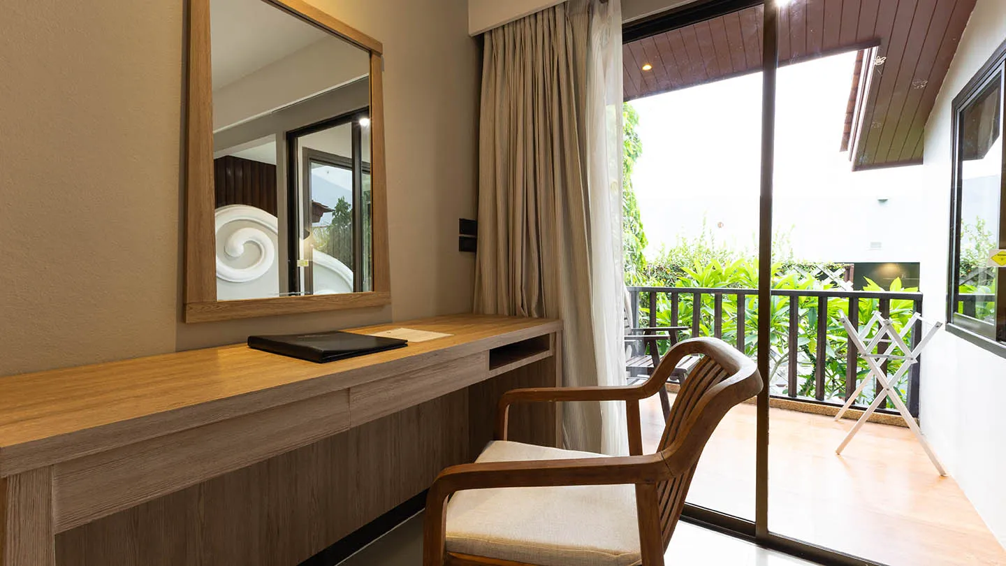 A wooden desk and chair with a framed mirror are positioned by a glass sliding door to a private balcony, featuring warm wood finishes and cream-colored walls bathed in natural light
