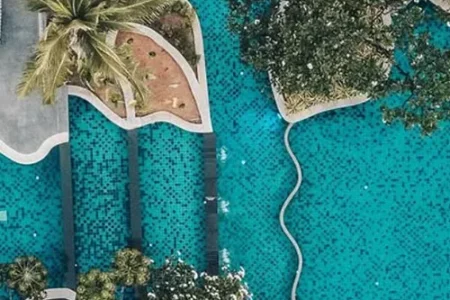 The resort showcases a cascading double-level pool amid lush greenery, sun loungers, and scenic sea views from above