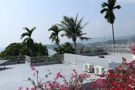 A scenic beach resort view of the Andaman Sea, framed by vibrant flowers, lush greenery, palm trees, and a light-colored building with a terrace
