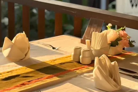 A table with an earthy-toned runner is set with plates, utensils, folded napkins, and a centerpiece of condiments and a small floral arrangement